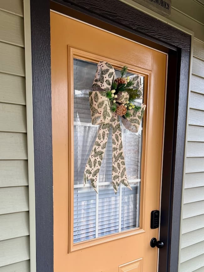 A tan door with a glass window panel is decorated with a large beige bow featuring green leaf patterns, pinecones, and small pink flowers. Framed by dark brown trim, the door has a keypad lock and is surrounded by light gray siding, adding charming lakehouse decor to the scene.