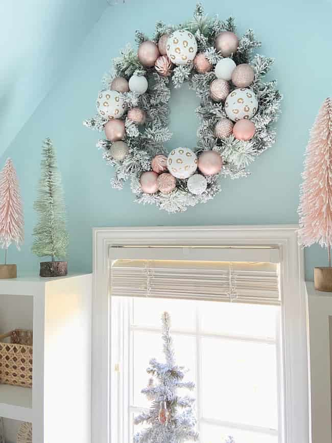 A festive holiday wreath with white and pink ornaments hangs on a light blue wall above a window. For Christmas theme ideas for decorating, flocked mini trees in pink and green can be displayed on the white shelves beside the window.