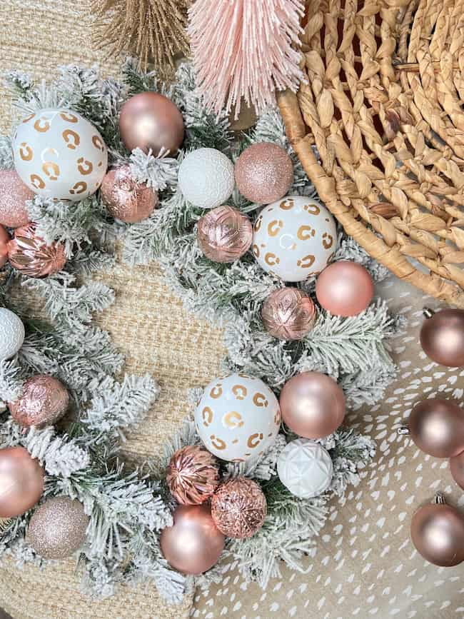 A holiday wreath with white and pink ornaments, featuring a leopard print design, is laid on textured fabric. Embracing Christmas theme ideas for decorating, the wreath is adorned with gold and pink tassels and sits gracefully near a woven basket.