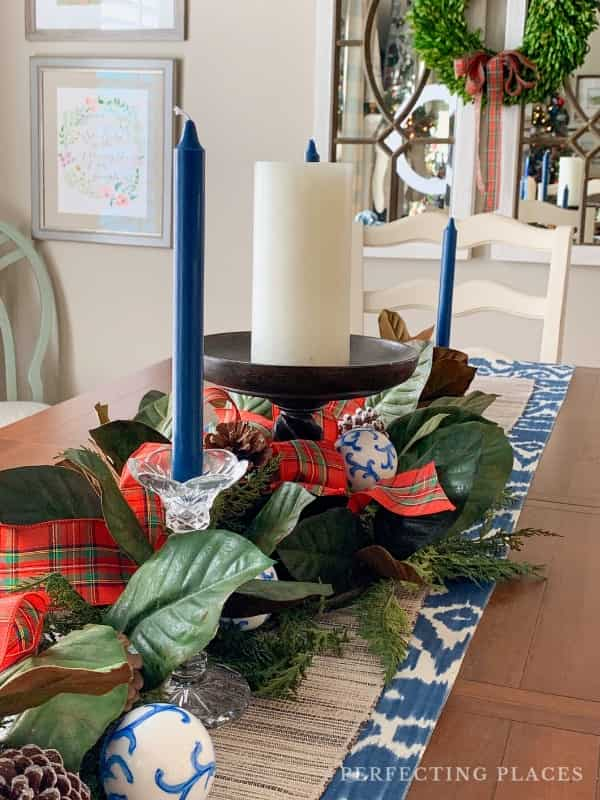 A dining table decorated with a holiday centerpiece embodies Christmas theme ideas for decorating, featuring dark blue taper candles, a white pillar candle, pinecones, green leaves, plaid ribbons, and blue and white ornaments on a patterned runner. Wall art and a wreath add charm in the background.