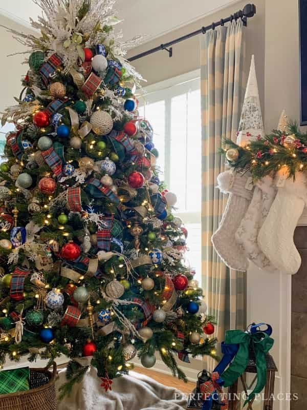 A beautifully decorated Christmas tree with colorful ornaments and ribbons stands by a window, offering inspiration for Christmas theme ideas for decorating. Two white stockings hang on the mantel, with wrapped gifts placed nearby, creating a cozy and festive holiday ambiance.