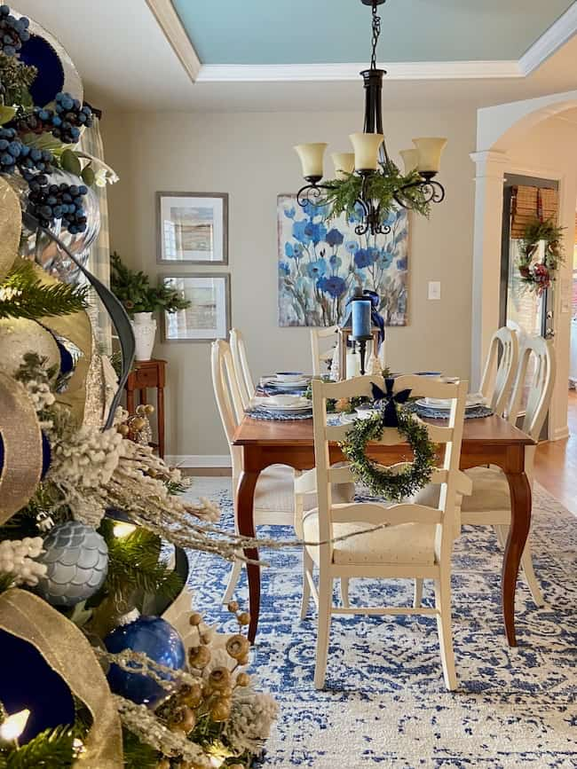 A dining room decorated with festive Christmas theme ideas for decorating, featuring a table set for dinner in blue and white accents. A Christmas tree with blue ornaments stands proudly in the foreground. A chandelier hangs above the table, while a wreath adds charm to a chair.