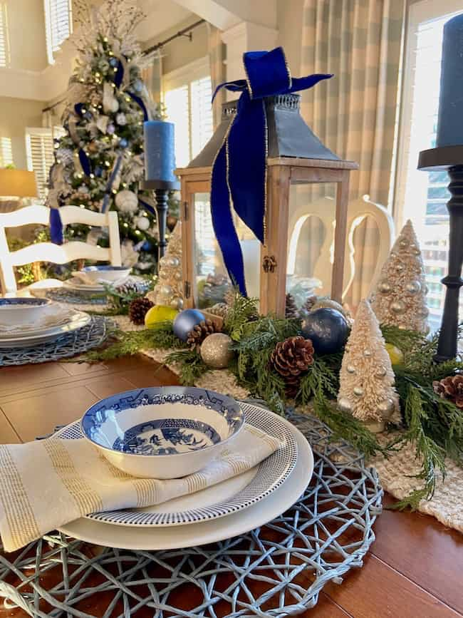 A festive dining table showcases Christmas theme ideas for decorating, featuring a blue and white bowl on a plate atop a woven placemat. The centerpiece includes a wooden lantern with a blue ribbon, surrounded by pinecones, ornaments, and decorative trees, with a Christmas tree in the background.