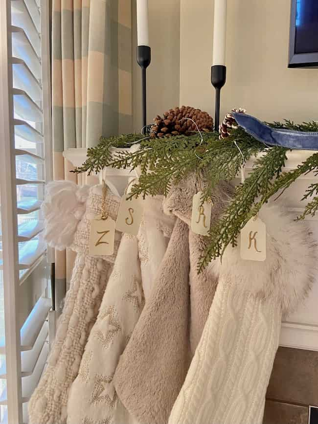 A cozy holiday mantel showcases charming Christmas theme ideas for decorating, with evergreen branches, pinecones, and three stockings labeled "Z," "S," and "K." Two candlesticks add warmth to the scene, while partially open window shutters complete the festive ambiance.