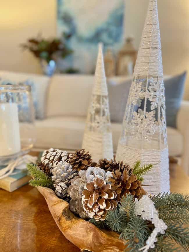 A cozy winter-themed living room exudes Christmas theme ideas for decorating, featuring cone-shaped trees, snow-dusted pine cones, and evergreen branches in a wooden bowl on the table. In the background, a blurred couch stands beside a blue abstract painting.