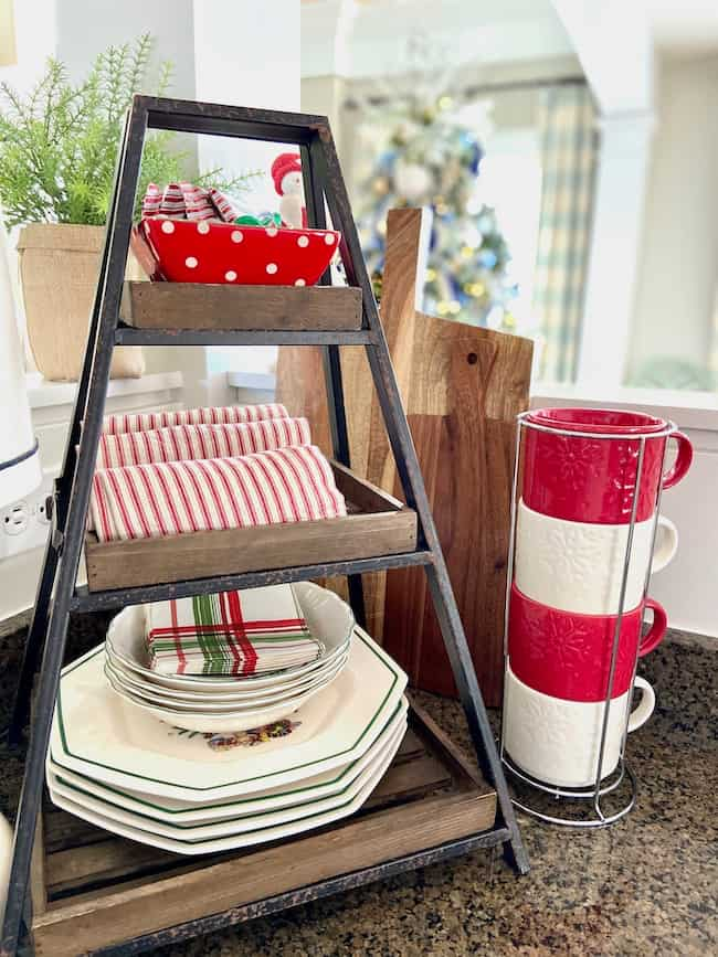 A kitchen counter adorned in a Christmas theme features a charming wooden tiered shelf with festive napkins and plates. Beside it, red and white mugs are neatly stacked in a holder, while a potted plant adds greenery to the holiday décor.