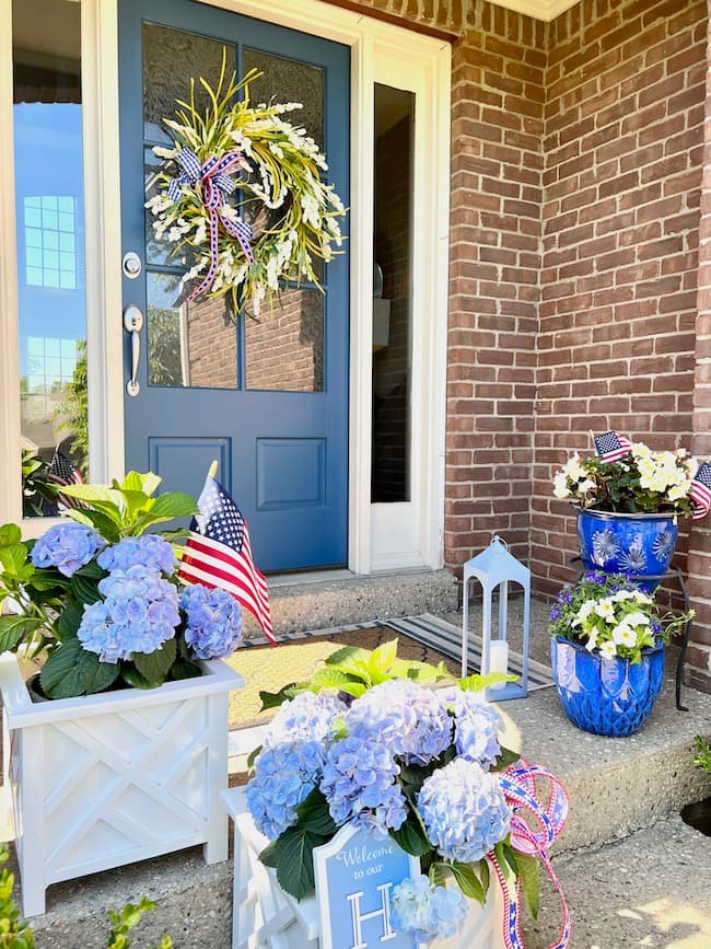 Memorial Day patriotic pots with blue hydrangeas in white flower boxes, blue flower pots with white flowers, and blue front door with patriotic wreath
