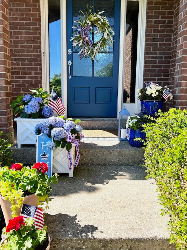 Memorial Day patriotic pots with blue hydrangeas in white flower boxes, blue flower pots with white flowers, red geraniums in terracotta pots, and blue front door with patriotic wreath