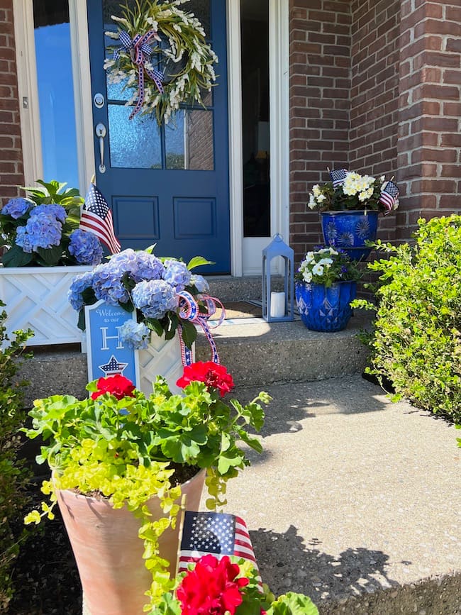 Memorial Day patriotic pots with blue hydrangeas in white flower boxes, blue flower pots with white flowers, red geraniums in terracotta pots, and blue front door with patriotic wreath