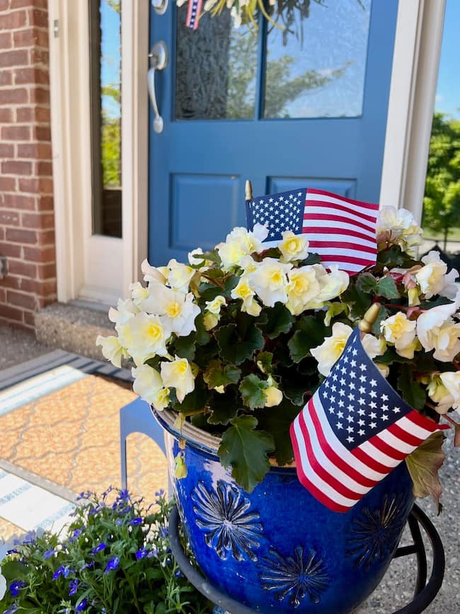 Small American flags in blue flower pot with white begonia flowers on the front porch
