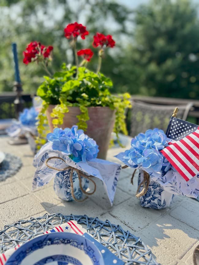 Memorial Day Tablescape with Potted red geraniums and blue and white napkin-wrapped mason jars with hydrangeas