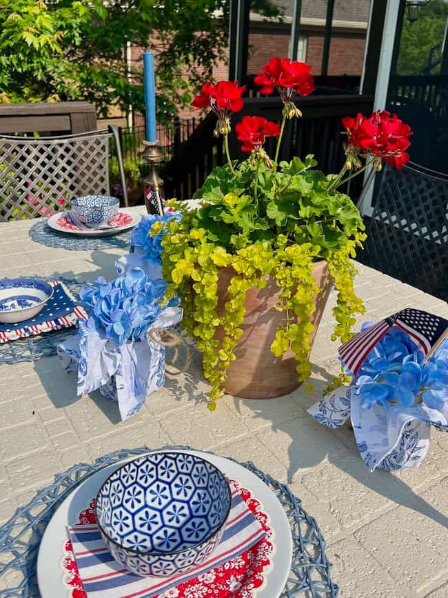 Memorial Day Tablescape with Potted red geraniums and blue and white napkin-wrapped mason jars with hydrangeas