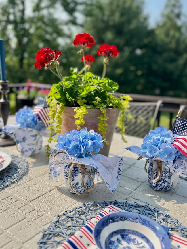 Memorial Day Tablescape with Potted red geraniums and blue and white napkin-wrapped mason jars with hydrangeas