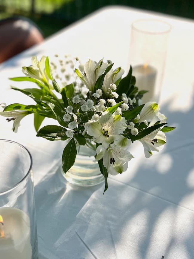 Backyard engagement party centerpiece with simple small arrangement of white alstroemeria and baby's breath with pillar candles in cylinder vases.