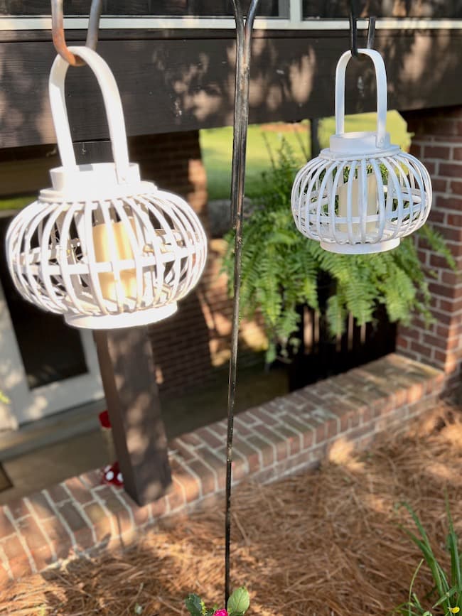 Wooded white lanterns with faux pillar candles hanging from shepherds hooks outdoors.