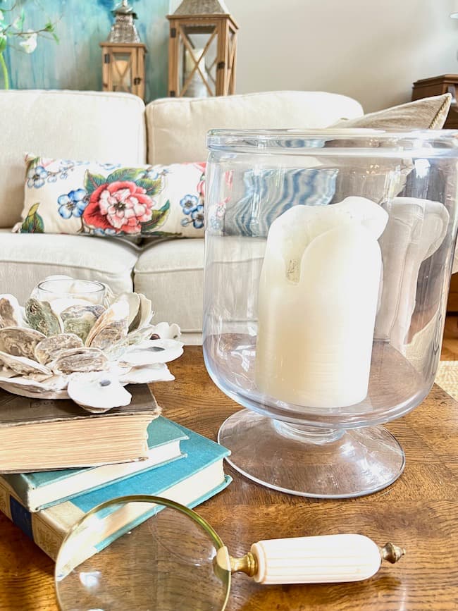 Textural elements on coffee table -- oyster shell votive holder, stack of vintage books, magnifying glass, moss covered balls in woven basket.