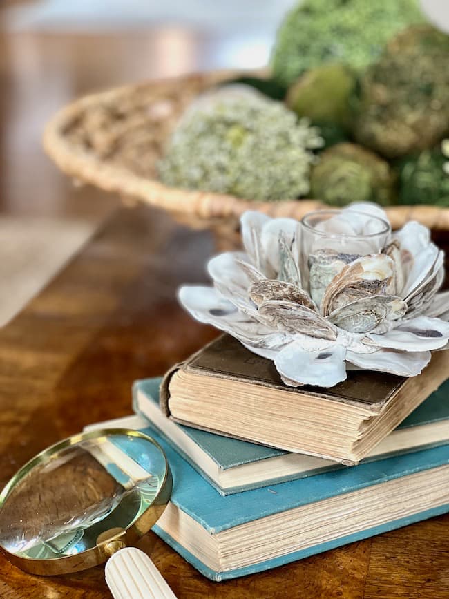 Textural elements on coffee table -- oyster shell votive holder, stack of vintage books, magnifying glass, moss covered balls in woven basket.