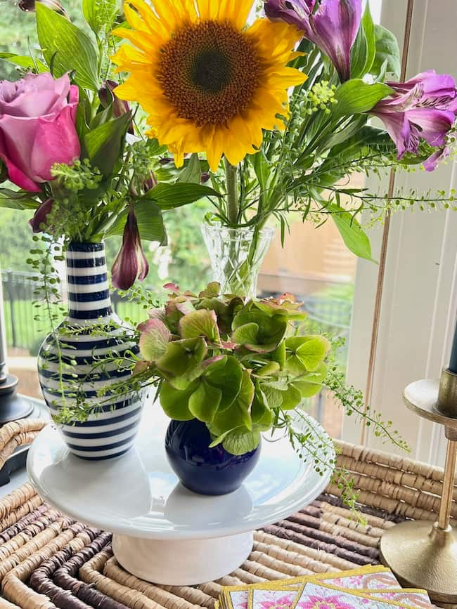 Mini bud vas arrangements on a white pedestal featuring sunflowers, pink roses, and hydrangea bloom