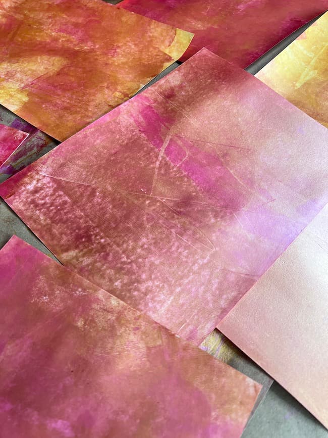 Abstract art pieces with a mix of pink, orange, and yellow hues are laid out on a flat surface. Alongside them, a DIY paper leaf garland for fall adds an extra touch of seasonal charm. The artwork features a blend of textures and colors, creating a vibrant and dynamic visual effect.