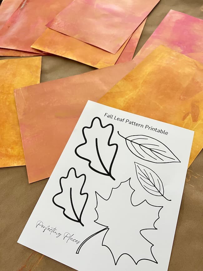 A set of colorful orange and pink paper sheets are laid out on a table. In the center is a printable sheet labeled "Fall Leaf Pattern Printable," featuring black outlines of different leaf shapes, ready to be cut out for your DIY paper leaf garland for fall.