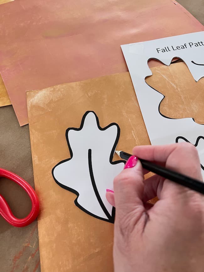 A person traces a fall leaf pattern onto a piece of orange paper using a black marker. The tracer template has multiple leaf shapes, perfect for creating a DIY paper leaf garland for fall. Nearby, red scissors await use, while the person's pink nail polish adds a pop of color.