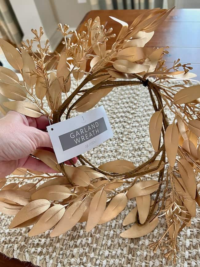 A hand holding a decorative garland wreath made of gold-colored leaves and small sprigs. The garland, reminiscent of a DIY paper leaf garland for fall, has a tag attached that reads "GARLAND WREATH." The wreath is displayed over a textured, beige tablecloth on a wooden table in a well-lit room.