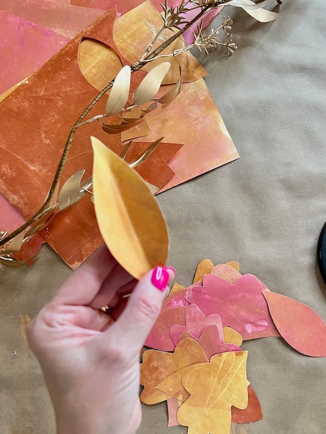 A hand with pink painted nails is holding a yellow-orange paper leaf, part of a DIY paper leaf garland for fall. More paper leaves in shades of yellow, orange, and pink are scattered on a brown surface, along with some bronze-colored foliage.