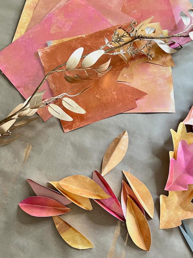 A collection of colorful paper sheets and paper cutouts arranged on a surface. The paper pieces resemble leaves in hues of pink, orange, yellow, and gold. A dried branch with leaves is also included, adding a natural element to this DIY paper leaf garland for fall setup.