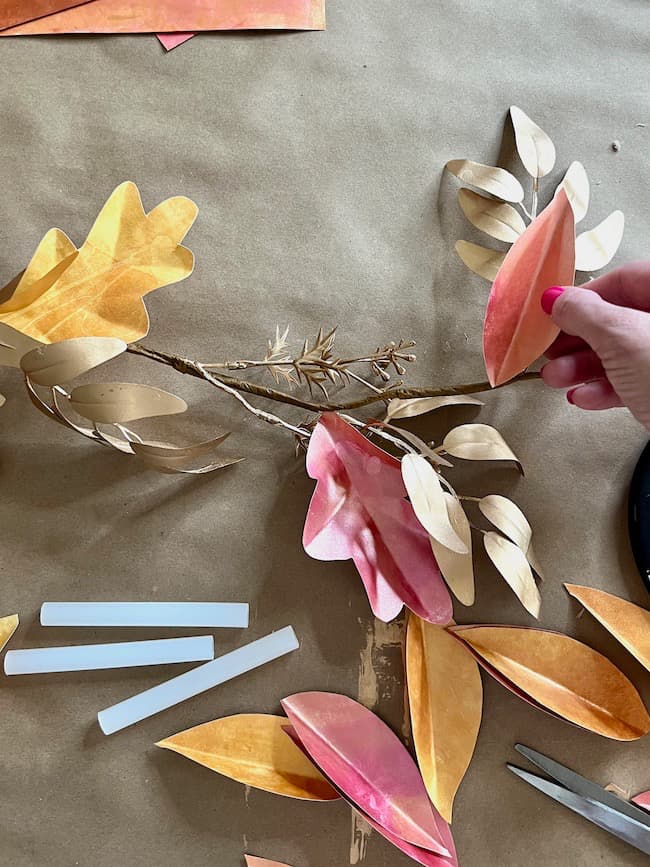 A person is holding a paper leaf next to a branch adorned with other colorful paper leaves in shades of pink, orange, and beige. Surrounding the branch are more paper leaves and white adhesive strips—the perfect setup for creating a DIY paper leaf garland for fall. The workspace has a brown paper covering.