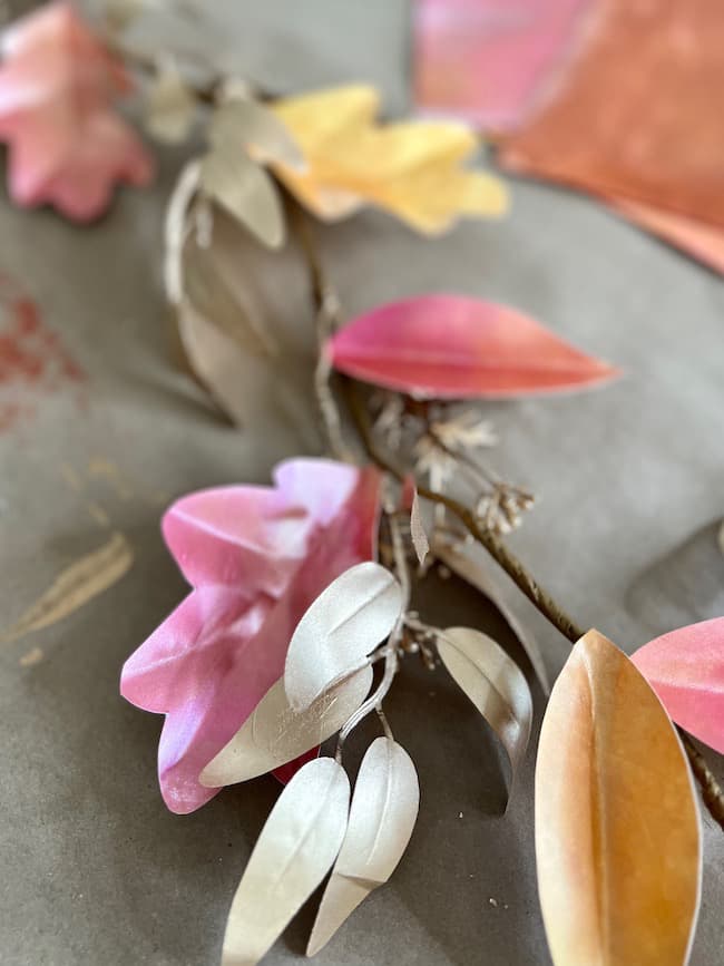 A section of a DIY paper leaf garland for fall features realistic, colorful paper leaves in shades of pink, yellow, orange, and beige arranged on a gray surface. The leaves have a matte finish and are delicately detailed, giving a natural and artistic appearance.