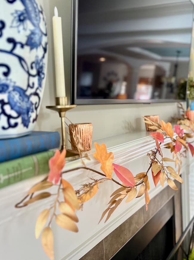 A close-up of a decorated mantel features a gold candlestick with a white candle, a blue and white vase, vintage books, and a DIY paper leaf garland for fall. The scene is set against a flat-screen TV above a fireplace, creating a cozy and festive atmosphere.