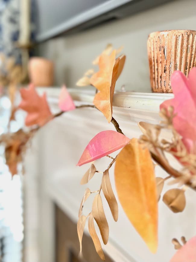 A DIY paper leaf garland for fall, made of vibrant orange, pink, and beige paper leaves, is draped over a white fireplace mantle. In the background, two terracotta-colored candle holders add a cozy touch to the scene.