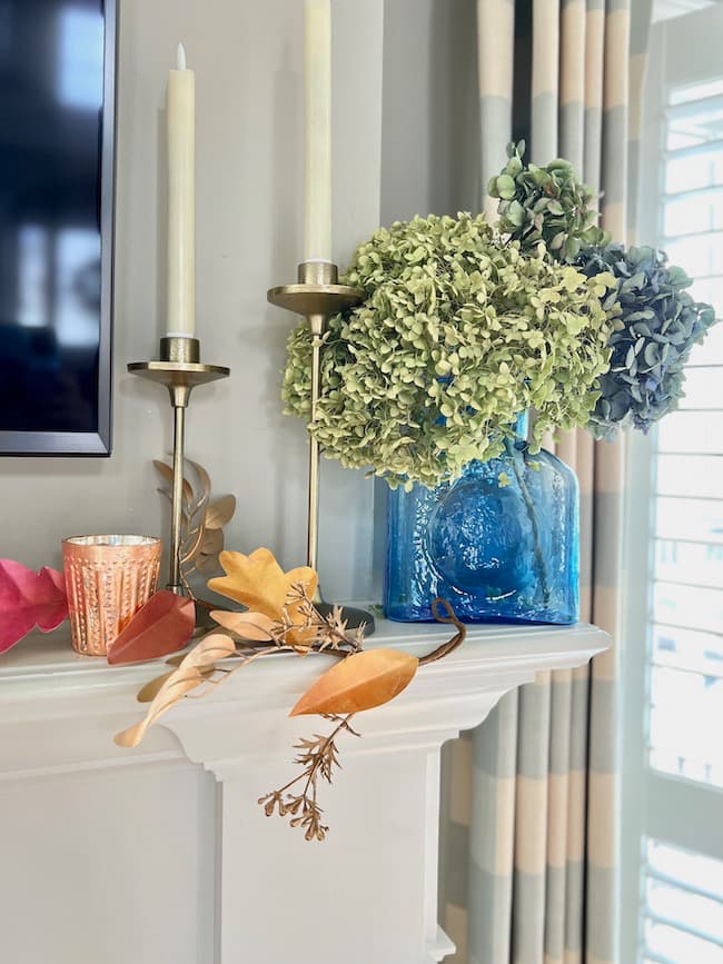 A white mantel is decorated with tall gold candlesticks holding cream candles, a blue glass vase filled with green and blue hydrangeas, a small copper votive candle holder, and autumn leaves scattered around, complemented by a DIY paper leaf garland for fall, with striped curtains and a shuttered window in the background.