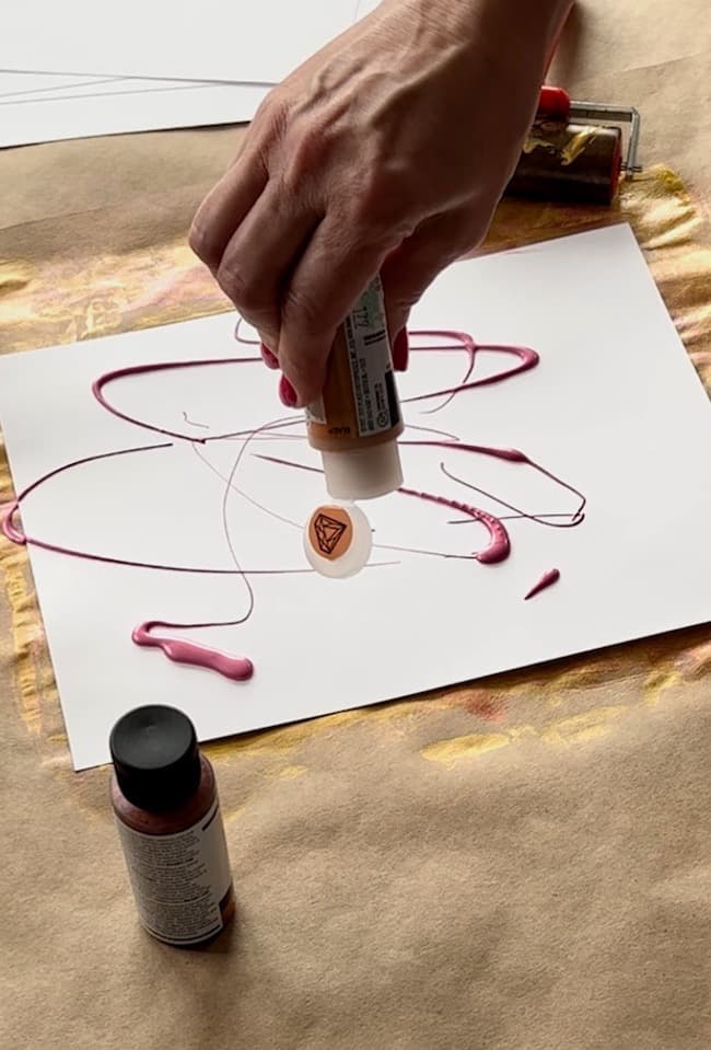 A hand is holding a paint container, squeezing pink paint onto a sheet of white paper with some abstract lines, perhaps for a DIY paper leaf garland for fall. Additional bottles of paint are on the table around the paper. The surface underneath appears to be a brown material.