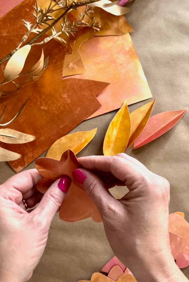 Two hands with red painted nails are seen holding a paper petal. The background features orange and yellow paper petals and leaves, along with a sprig of foliage, set on a brown surface. The scene suggests a DIY paper leaf garland for fall in progress.