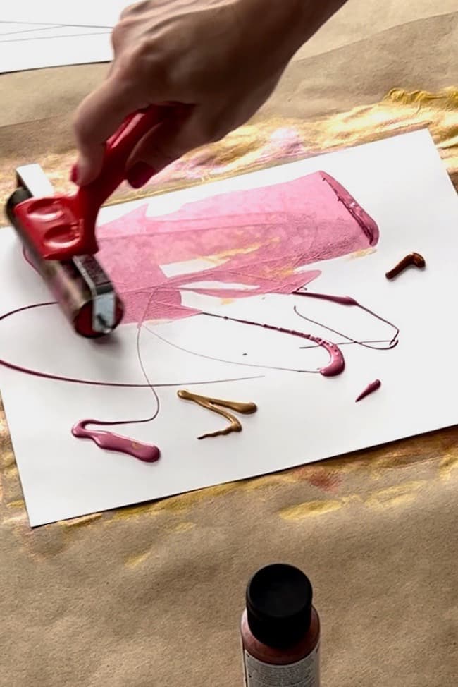 A person uses a red roller to spread pink paint over white paper. Various streaks and blobs of paint in pink and brown are also visible on the paper, which is lying on a brown surface. A bottle of paint is seen below the paper, likely for creating a DIY paper leaf garland for fall.