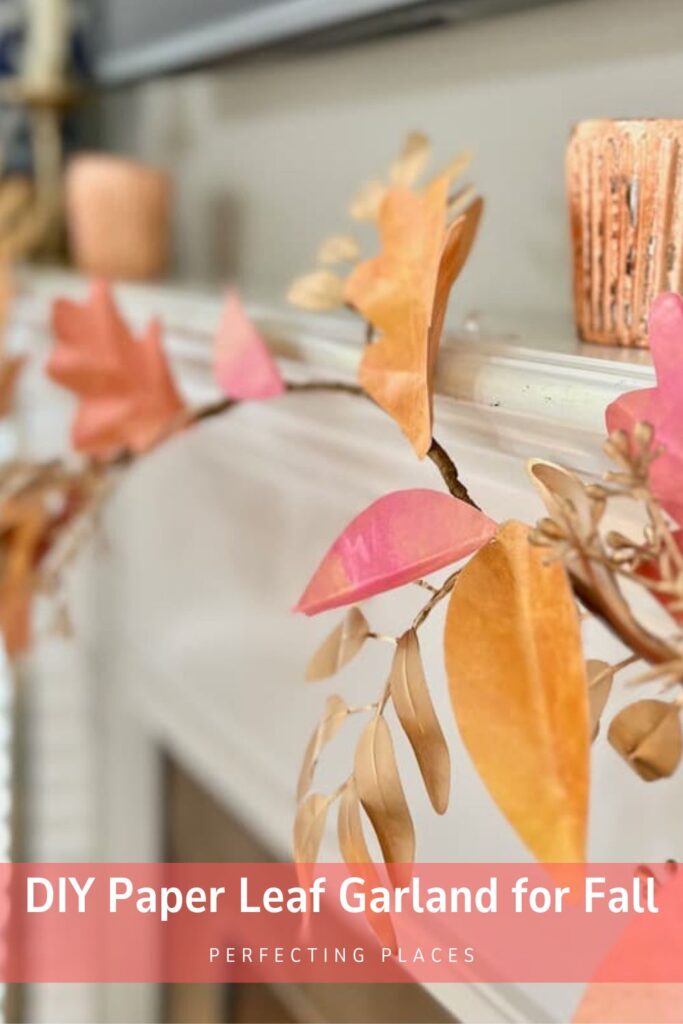 A close-up of a mantel decorated with a charming DIY paper leaf garland in autumn colors, featuring vibrant orange, pink, and gold leaves. A candleholder and various decor pieces grace the background. Text at the bottom reads "DIY Paper Leaf Garland for Fall - Perfecting Places.