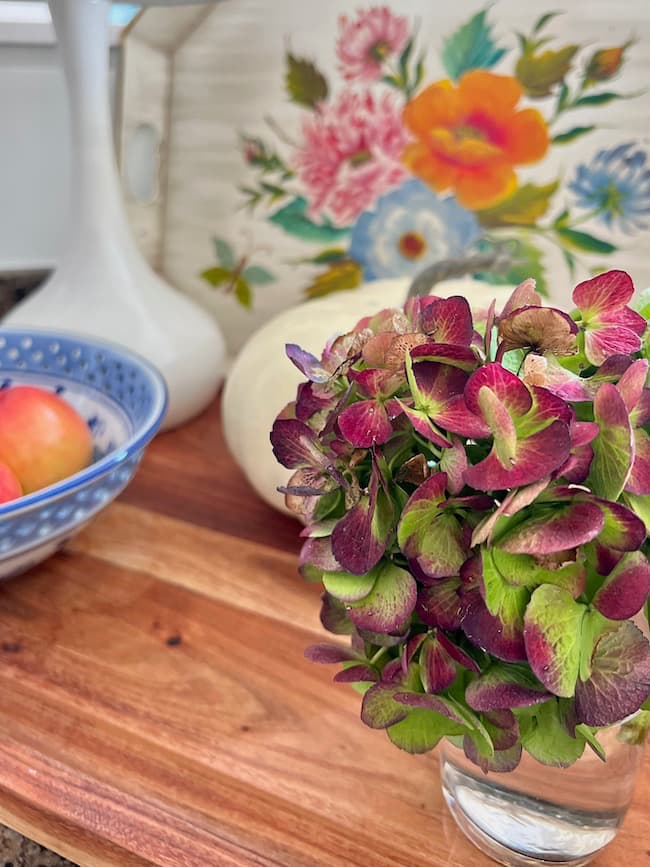 Fall Decor on Kitchen Countertops with a dried hydrangea bloom, vintage tole painted tray, blue and white bowl, red apples on a wooden cutting board.