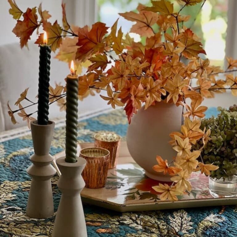 A cozy autumn table setting showcases budget-friendly fall kitchen decor ideas with two lit spiral candles in holders, a vase of vibrant leaves, and a floral-patterned tray. The textured tablecloth in green and blue adds depth, enhancing the warm, seasonal ambiance.