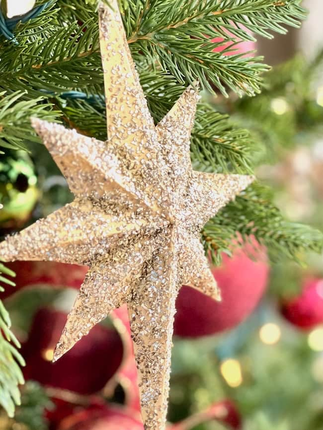 Close-up of a creative DIY glittery gold star ornament hanging on a Christmas tree. The star is surrounded by green pine needles and blurred red and gold baubles in the background.