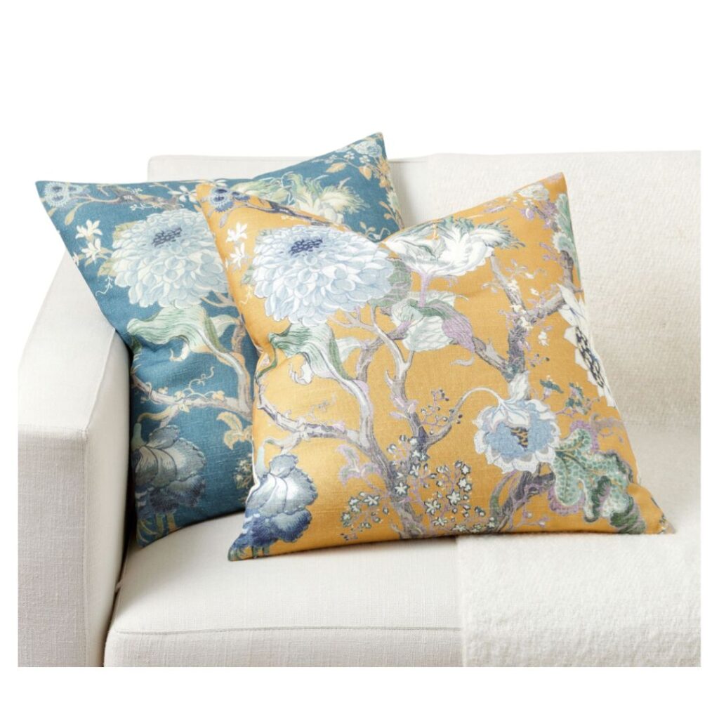 Two decorative pillows with floral patterns rest on a light-colored couch, perfect to decorate for spring. One pillow boasts a teal background, while the other showcases mustard yellow. Both feature intricate designs of leaves and flowers, adding a refreshing touch to your space.