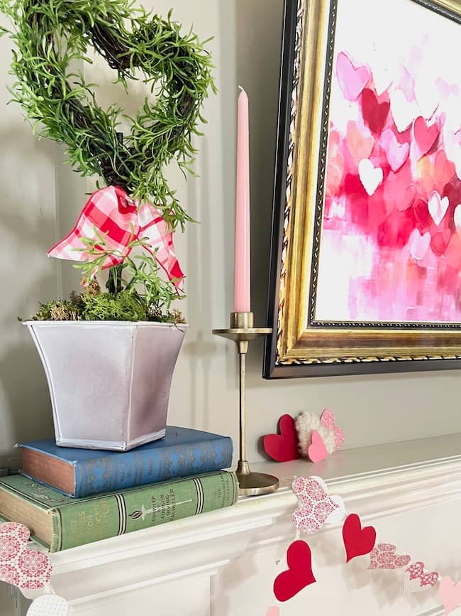 Valentine heart topiary on a stack of books on the mantel with pink candle in gold candleholder next to heart-themed digital TV art and paper heart garland