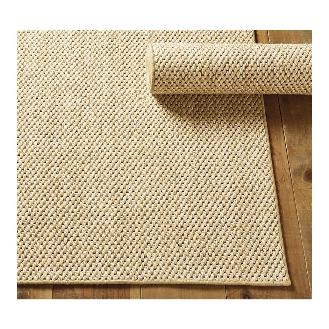 A beige woven rug, possibly a jute or sisal variety, is partially rolled on a wooden floor. The rug boasts a textured appearance with its thick, braided pattern, blending understated elegance with natural charm.