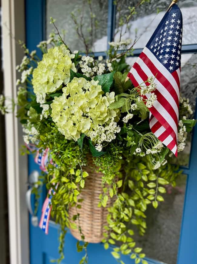 A woven basket filled with green and white flowers, greenery, and an American flag hangs on a blue door. Red, white, and blue ribbon accents this patriotic home decor arrangement, creating a festive tribute to America.