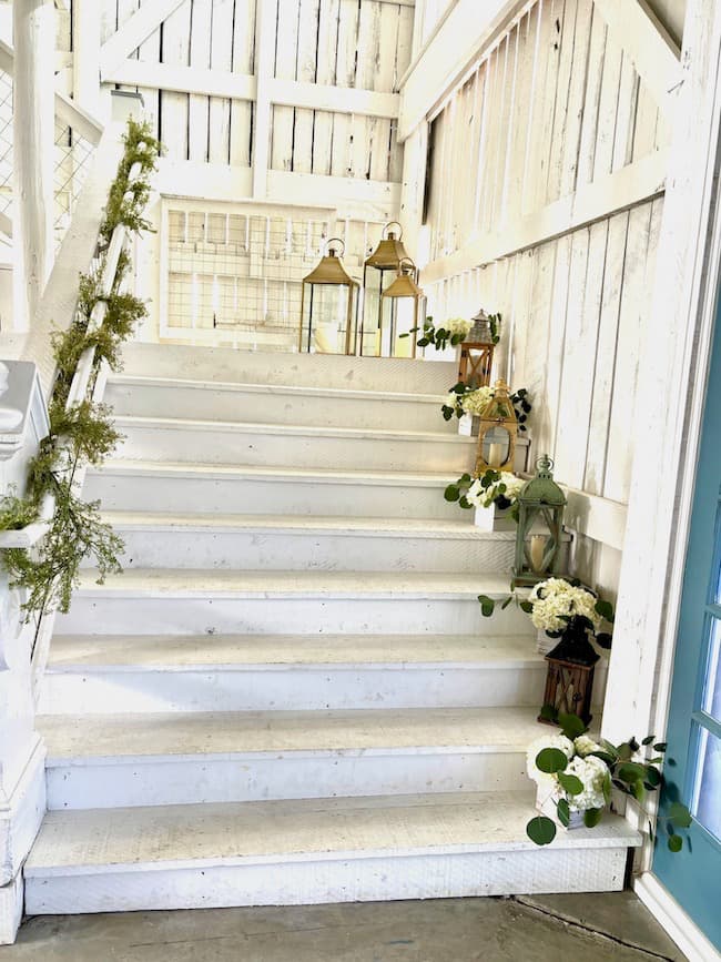 Large white staircase in white barn venue for wedding decorated with lanterns, hydrangea arrangements, and greenery