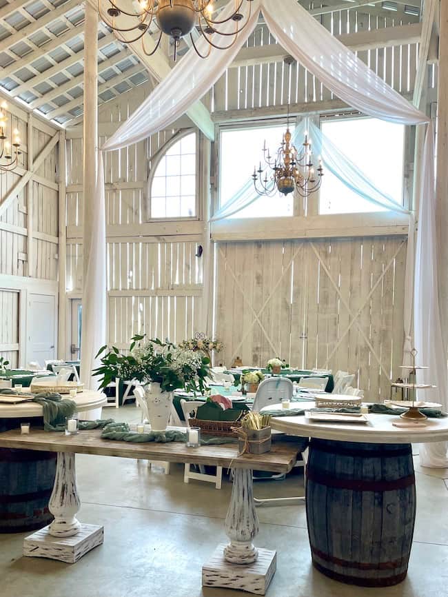 Wedding Venue, Zion Hill Events, for rehearsal dinner and wedding, featuring a white barn with white draping and large chandeliers. Tables decorating in deep green tablecloths with hydrangea and eucalyptus centerpieces -- Various wood tables, including bourbon barrel bases serve as the dessert tables.