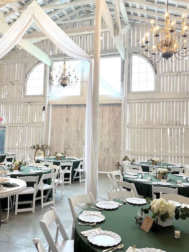 Wedding Venue, Zion Hill Events, for rehearsal dinner and wedding, featuring a white barn with white draping and large chandeliers. Tables decorating in deep green tablecloths with hydrangea and eucalyptus centerpieces.