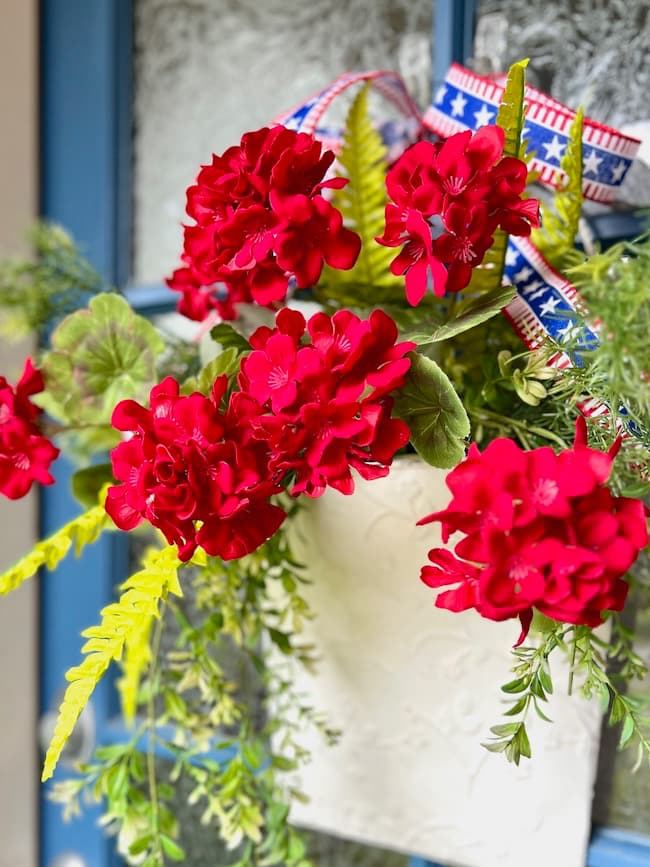 Patriotic door decoration featuring a white metal flower bucket with red geraniums and patriotic ribbon.