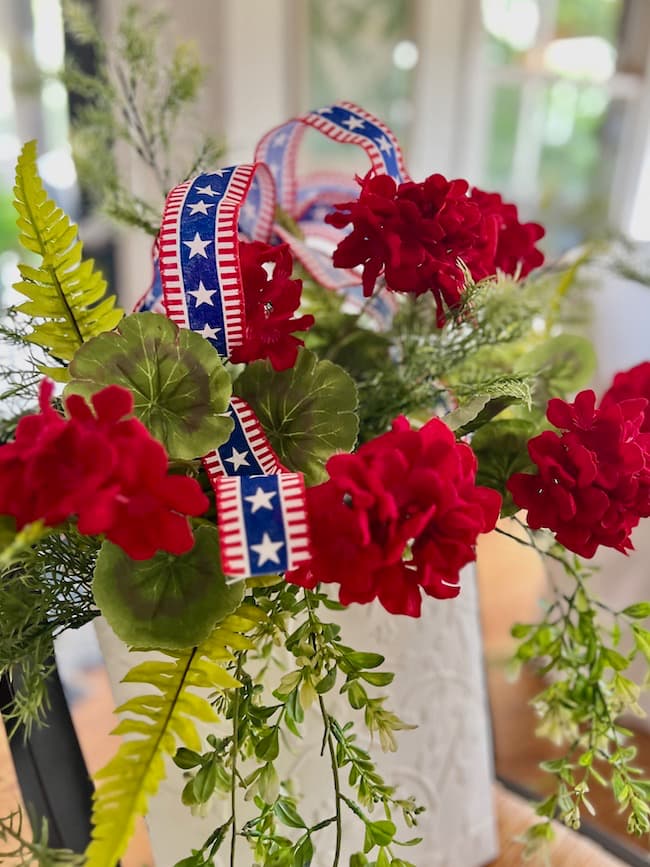 White flower bucket door hanger with faux fern greenery, faux red geraniums, and a red, white, and blue stars and strips ribbon.