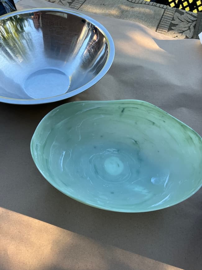 A large metal bowl and smaller plastic green and white swirled oval shaped bowl sit on a brown paper covered table waiting to be used as molds for DIY cement planters.